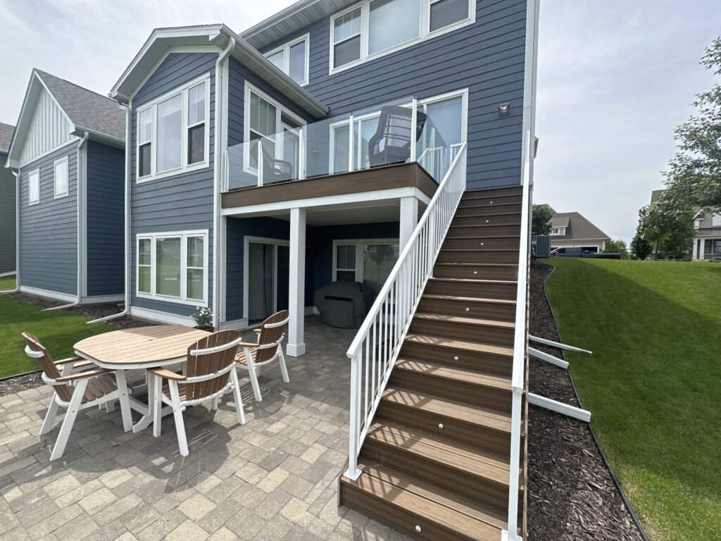 The back of a blue-sided home with a modern glass-railed upper deck and a lower paver patio furnished with a wooden dining table and chairs.