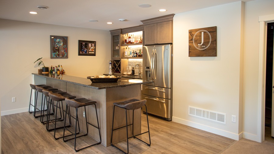 A sleek home bar with a granite countertop, backlit shelves displaying bottles, wine storage, a stainless steel refrigerator, and bar stools.
