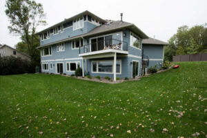 The rear view of a blue two-story house with a lower patio and an upper deck, situated on a sloped green lawn with scattered autumn leaves.