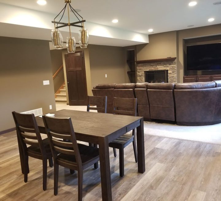 A finished basement featuring a dark wood dining table, a stone fireplace, a large curved sectional sofa for a home theater, and recessed lighting.