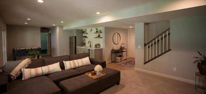 A cozy basement living room featuring a large brown sectional sofa, a wet bar with open shelving, and a view of the staircase leading to the upper level.