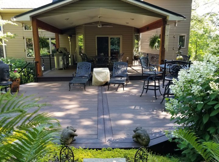 A shaded wooden patio with dark metal lounge chairs and tables, surrounded by lush greenery, ferns, and decorative stone statues.