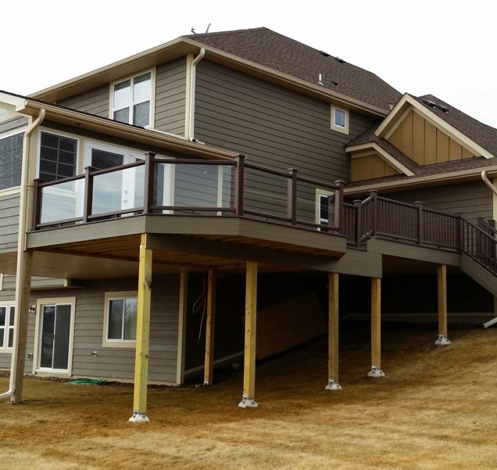 A substantial two-tiered deck with brown railings and stairs attached to a gray-sided house, situated on a sloped, dry grassy yard.