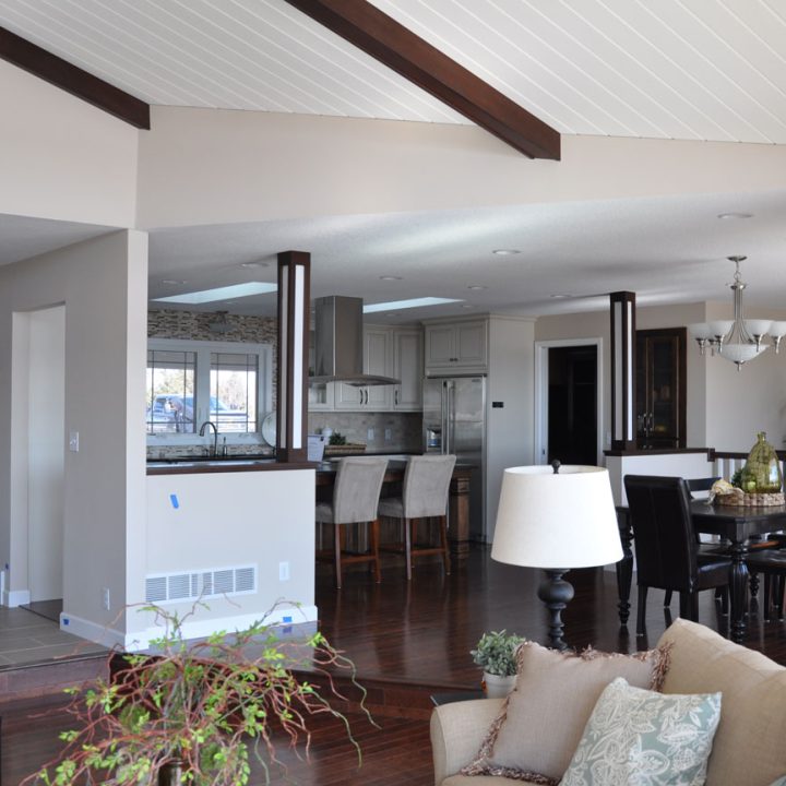 A modern open-concept living space with dark hardwood floors, exposed ceiling beams, and a view into the kitchen and dining area.