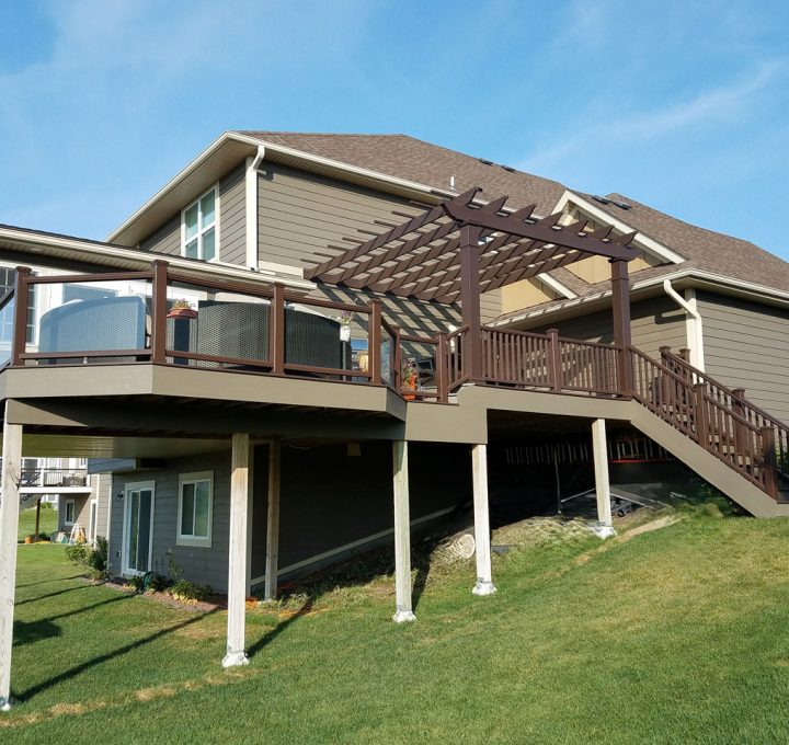 A large, modern deck with brown railings and a pergola attached to a gray-sided house on a sunny day.