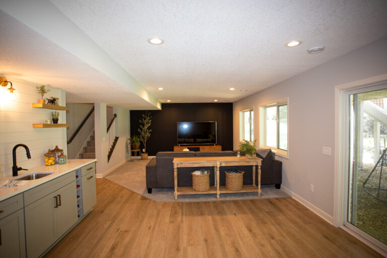 A finished basement featuring a wet bar with white cabinets, a large TV on a black accent wall, a gray sectional sofa, and a sliding glass door to the backyard.