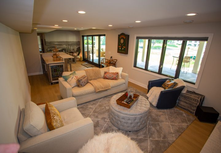 A cozy basement living room featuring a light-colored sectional sofa, round tufted ottomans, a dartboard on the wall, and large windows overlooking a patio.