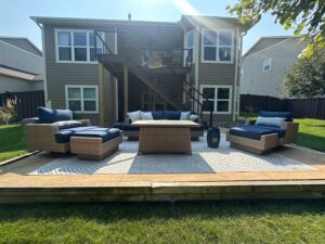 A lower-level patio with wicker furniture and a fire pit table on a patterned rug, situated in front of a gray home with a modern upper deck and stairs.