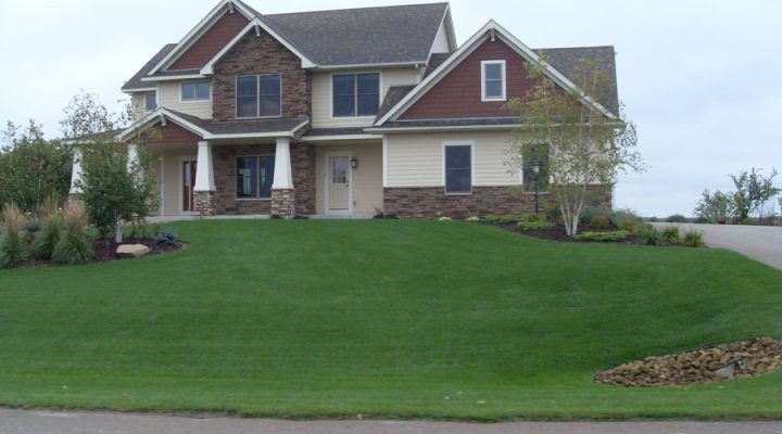 The front view of a two-story home featuring red and beige siding, stone accents, and a large, well-manicured green lawn under an overcast sky.