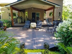 A shaded wooden patio with dark metal lounge chairs and tables, surrounded by lush greenery, ferns, and decorative stone statues.