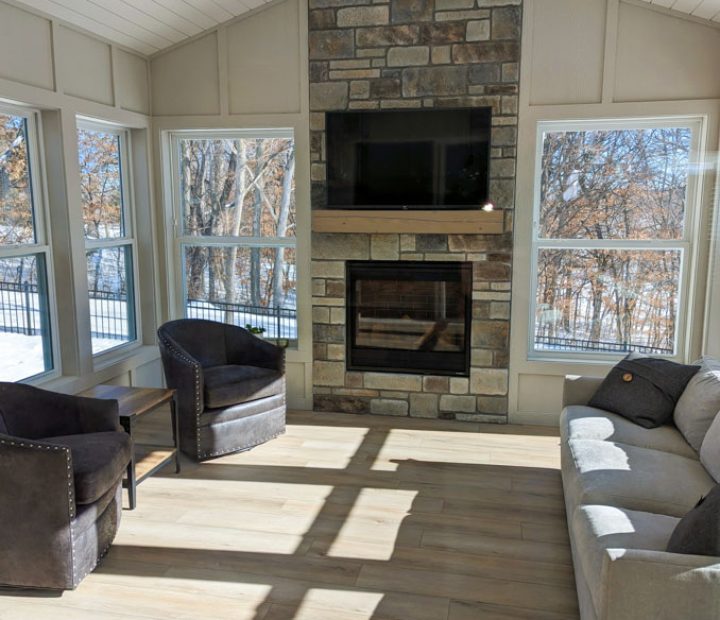 A cozy sunroom featuring a stone fireplace with a mounted TV, light wood floors, white shiplap ceiling, and large windows overlooking a snowy yard.
