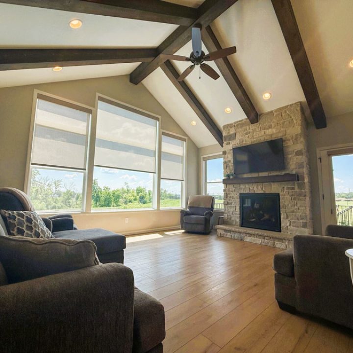 A spacious living room featuring a vaulted ceiling with exposed beams, a stone fireplace, hardwood floors, and large windows offering a view of the outdoors.