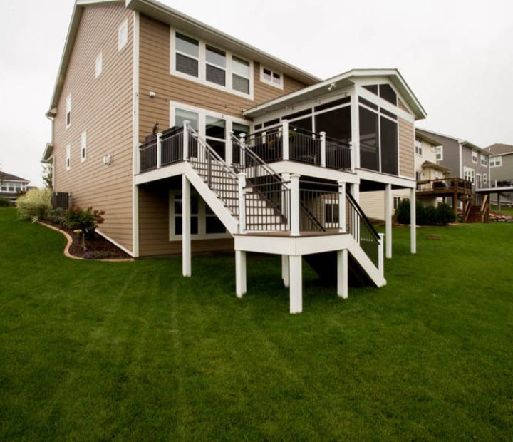 A two-tiered deck with black railings attached to a beige-sided house, situated on a landscaped yard with mulch and small plants.
