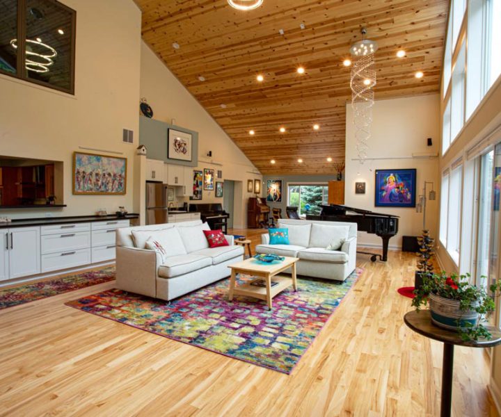 A bright, open-concept living room with a high vaulted wood ceiling, hardwood floors, white sofas, a colorful area rug, and a view into the kitchen.