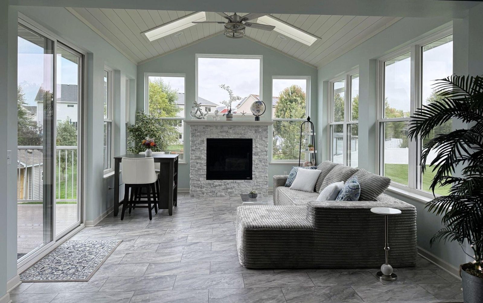 Interior of a 14 by 16 foot four-season porch in Rosemount, MN with a vaulted tongue-and-groove ceiling, skylights, ceiling fan, stone gas fireplace and walls of windows